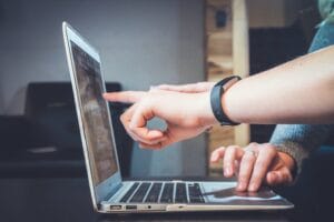 Close up image of a laptop, the hand of one person uses the track pad, the hand of another person point to the screen.