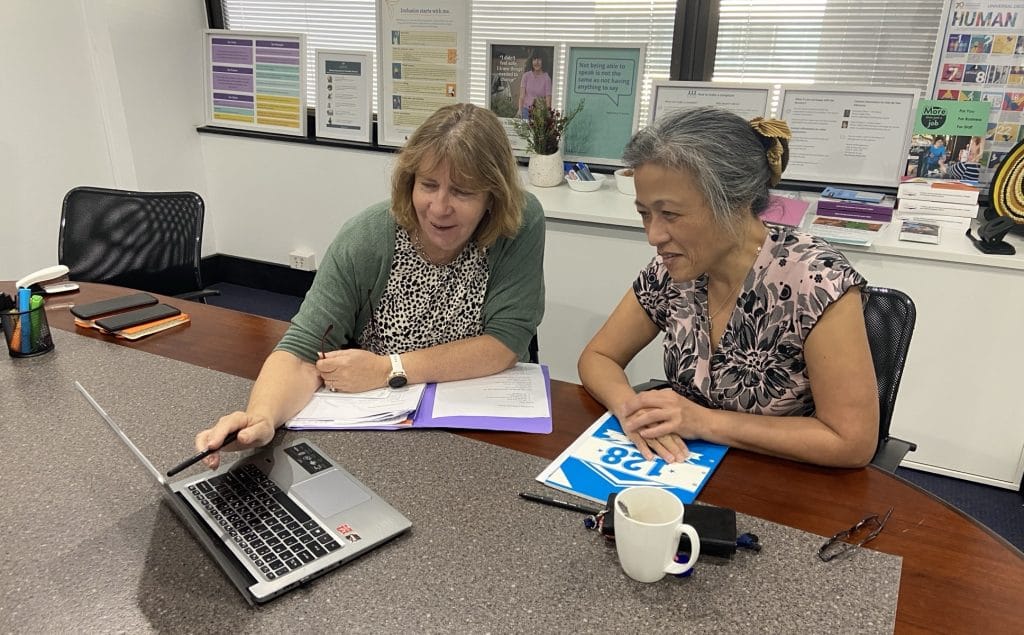 Two women sitting at a table. They are having a meeting. One woman is pointing at the screen of a laptop.