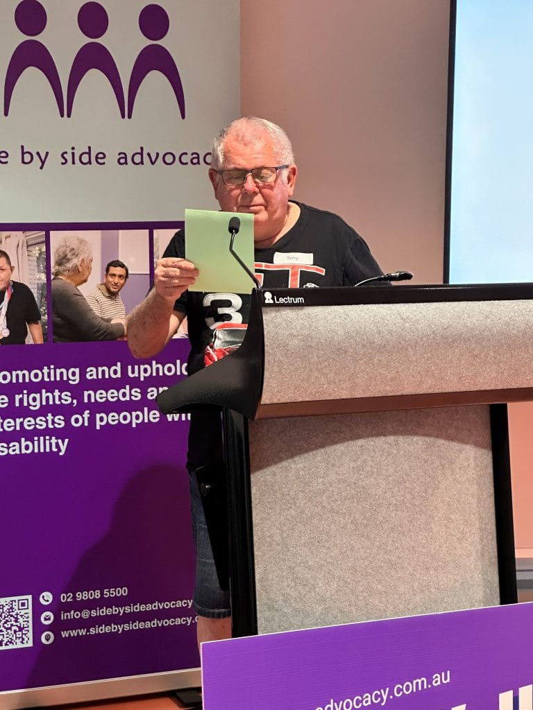 An older man is standing behind a lecturn. He is reading from a piece of paper