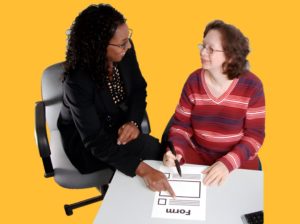 A woman in a black suit is helping a woman in a red striped shirt fill in a form.