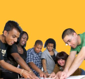 Six people are gathered around a table, pointing at a large sheet of paper together.
