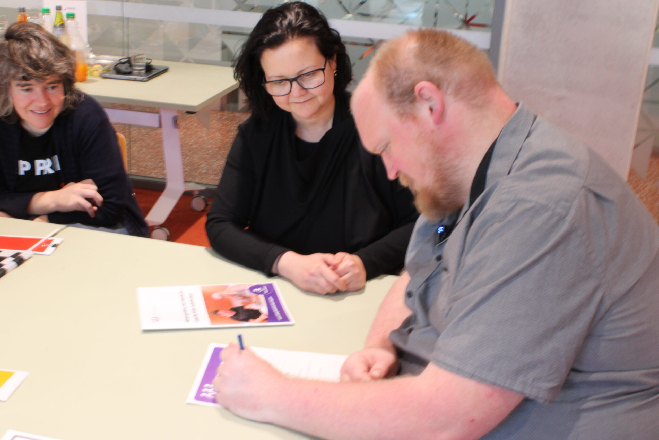 A man sits at a table signing a document while two women look on.